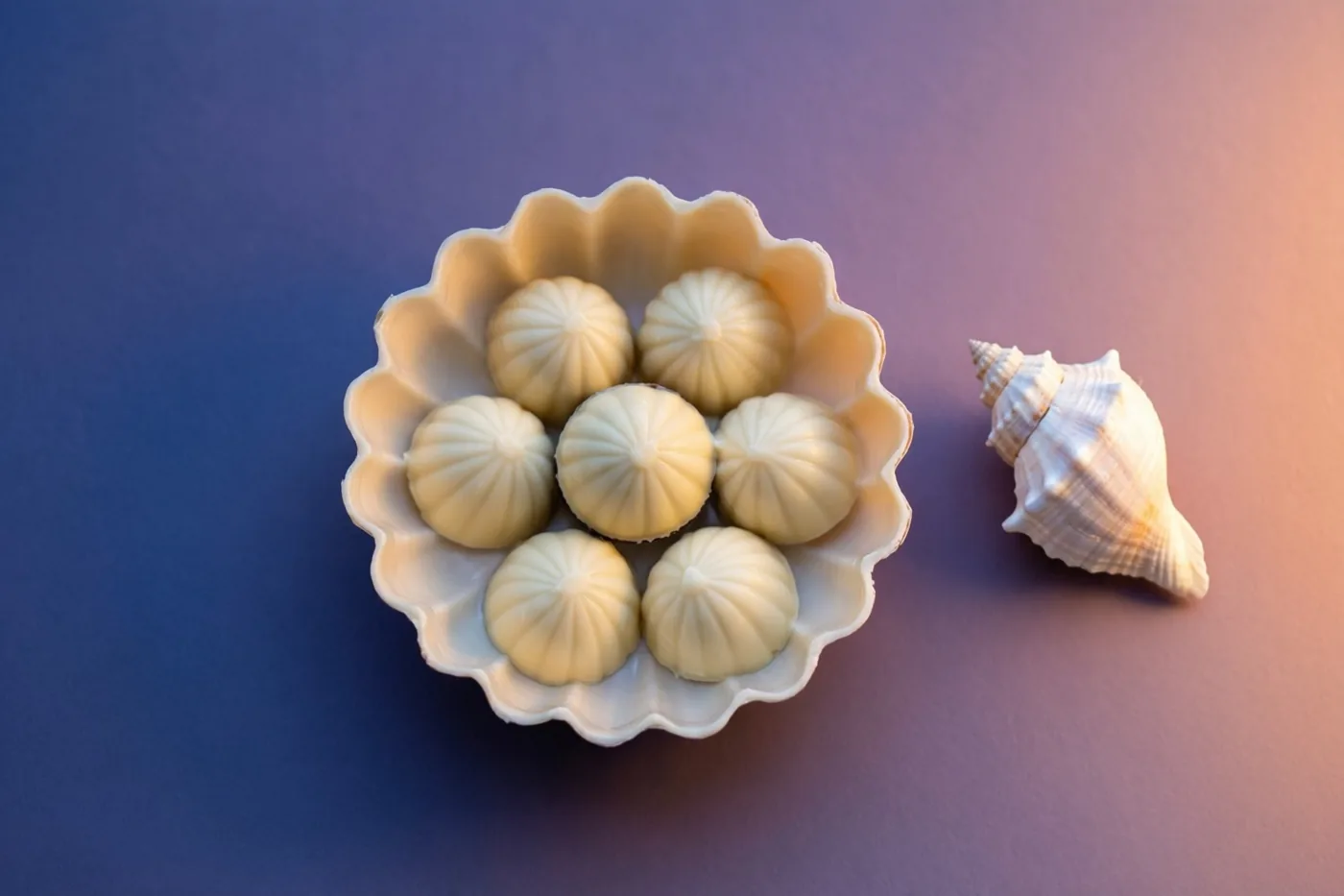 Zeven witte chocoladepralines liggen in een witte kom met geschulpte rand naast een kleine witte schelp, tegen een achtergrond die van paars naar blauw verloopt.