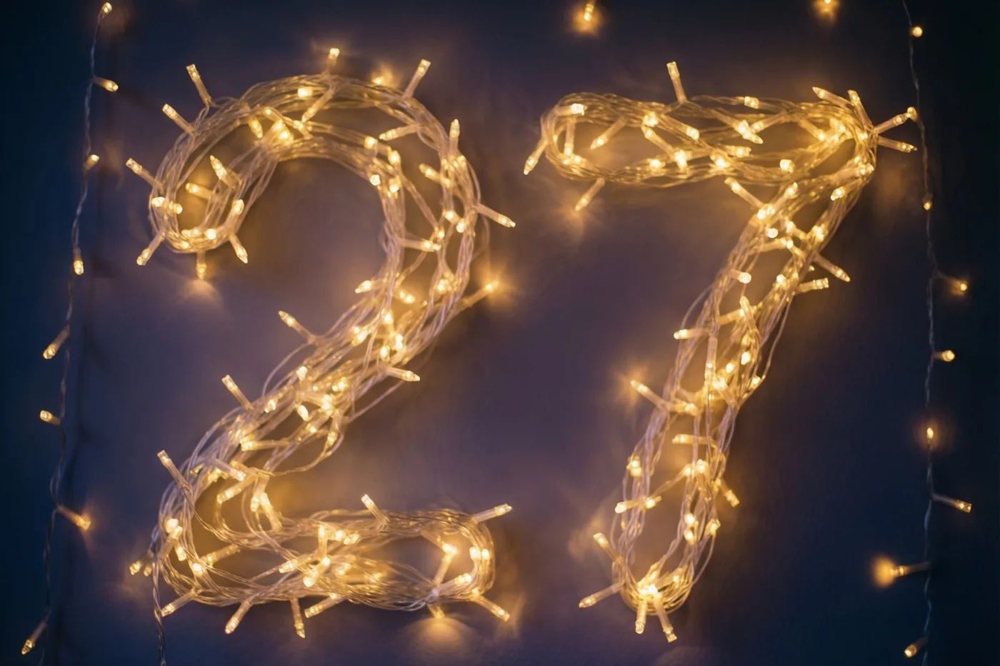 Warme lichtslingers vormen het getal 27 tegen een donkere muur, met zachte onscherpe lichtvlekken eromheen.