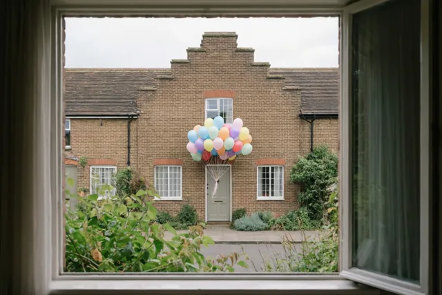Door een open raam is een bakstenen rijhuis met trapgevel en kleurrijke ballonnen aan de voordeur te zien, met groen op de voorgrond onder een bewolkte hemel.