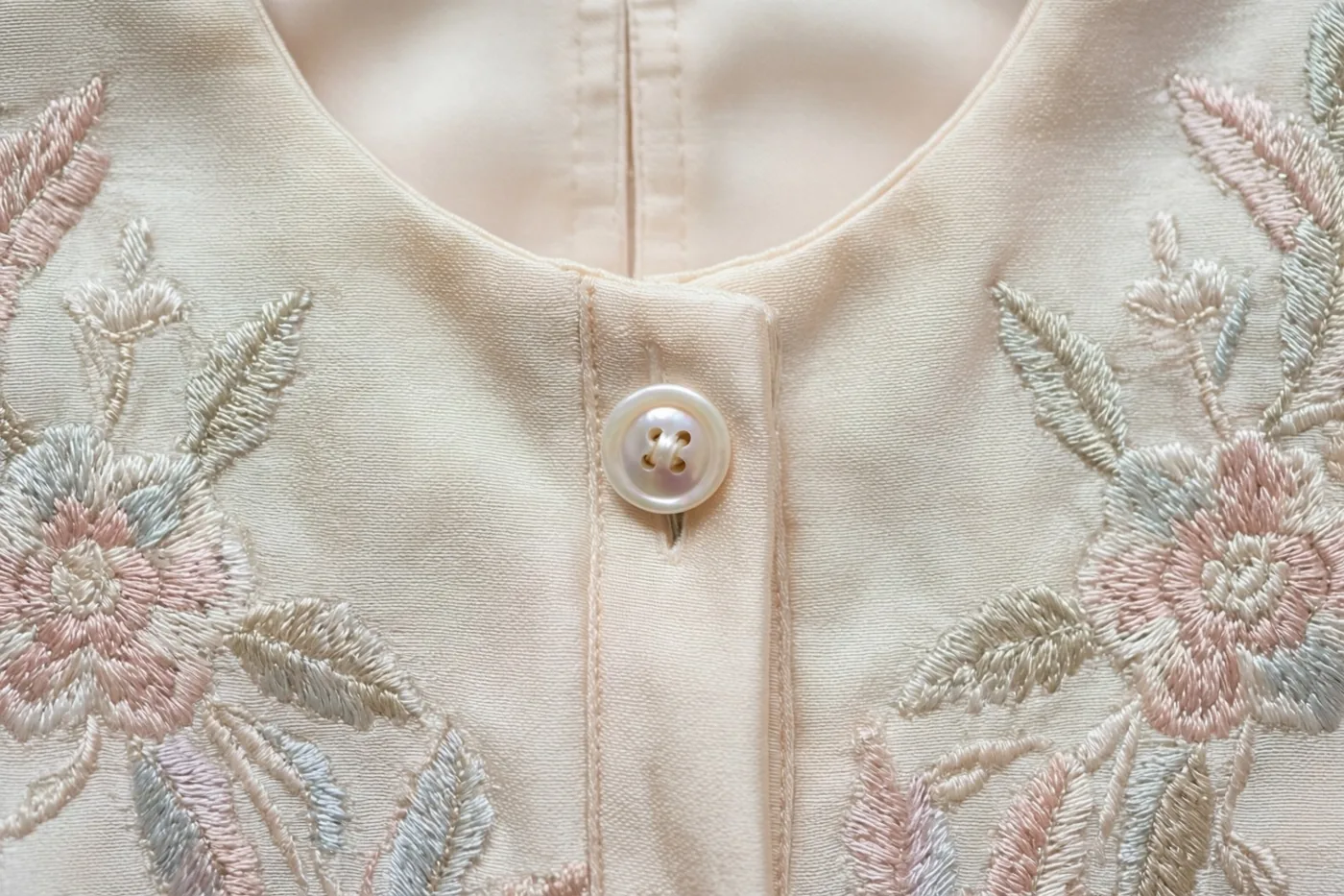 Een detailfoto toont een zijden doopkleed met fijn bloemenborduurwerk in pasteltinten en een klein parelmoeren knoopje aan de hals.