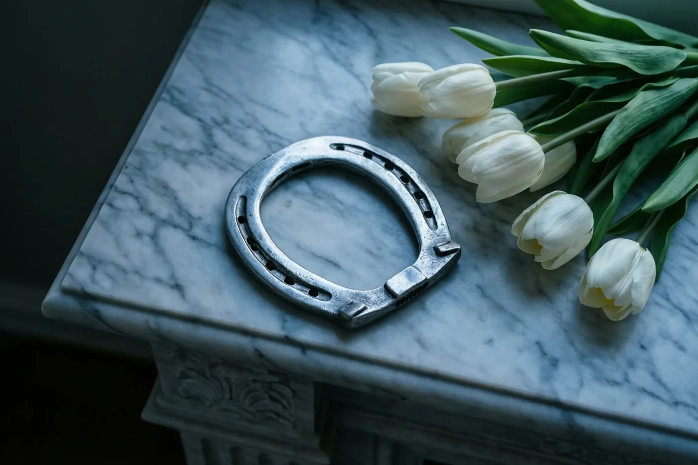 Een zilveren hoefijzer ligt op een witte marmeren schouw naast een boeket witte tulpen in een elegant, schemerig interieur.