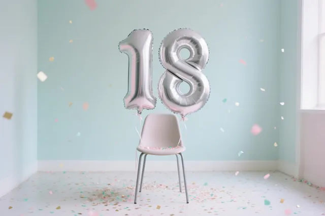 Twee grote zilveren folieballonnen vormen 18 aan een lichtroze stoel in een kamer met mintgroene muren, met roze, gouden en mintgroene confetti op de vloer en in de lucht.