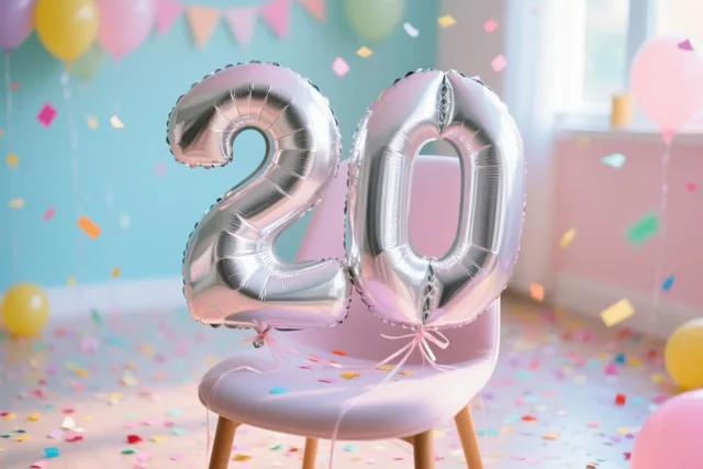 Zilveren folieballonnen vormen het getal 20 aan een roze stoel, met kleurrijke confetti, pastelballonnen en vlaggetjes in een lichte kamer.