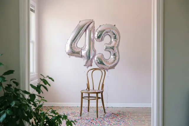 Zilveren folieballonnen vormen 43 aan een houten stoel in een lichte kamer met pastelkleurige muren en confetti op de vloer.