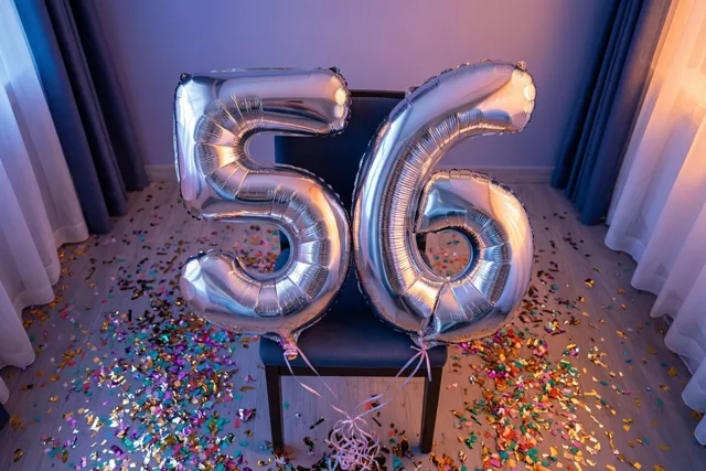 Twee grote zilveren folieballonnen vormen 56 aan een donkere stoel, met confetti op de vloer en zacht paars en warm oranje licht in de kamer.