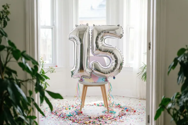 Zilveren folieballonnen vormen 15 aan een stoel in een lichte witte kamer met confetti op de vloer, omringd door groene planten en ramen.