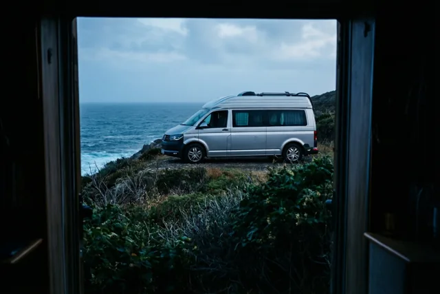 Door een open deuropening is een zilverkleurige camper te zien op een met gras begroeide klif boven de zee onder een bewolkte hemel.