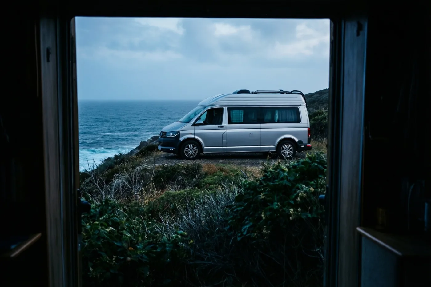 Door een open deuropening is een zilverkleurige camper te zien op een met gras begroeide klif boven de zee onder een bewolkte hemel.