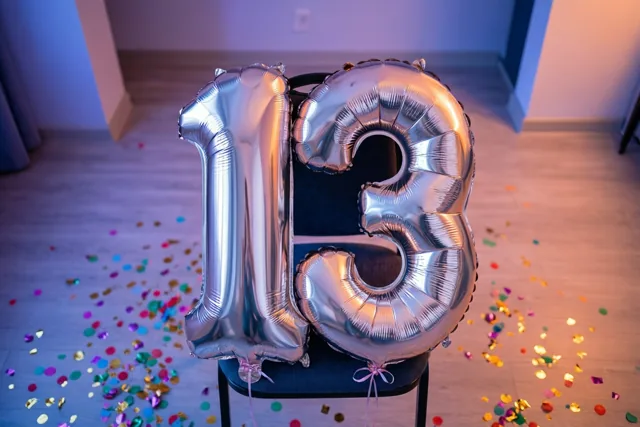 Twee grote zilveren folieballonnen vormen 13 aan een zwarte stoel, met kleurrijke confetti op de vloer en zacht blauw en warm oranje licht in de kamer.