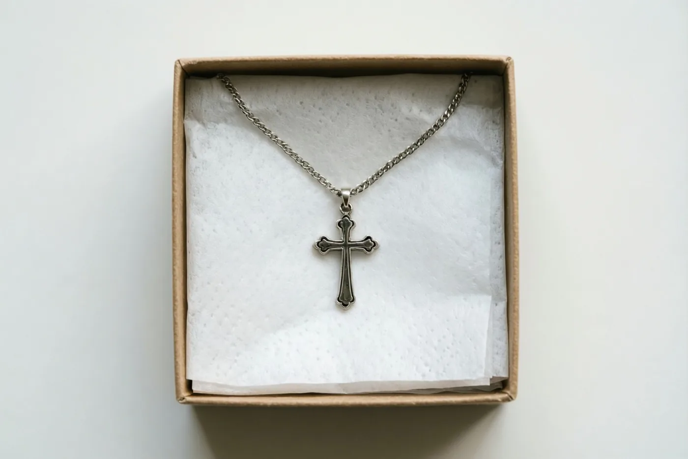 Een zilveren halsketting met een kruis ligt op wit zijdepapier in een open kartonnen geschenkdoos tegen een witte achtergrond.