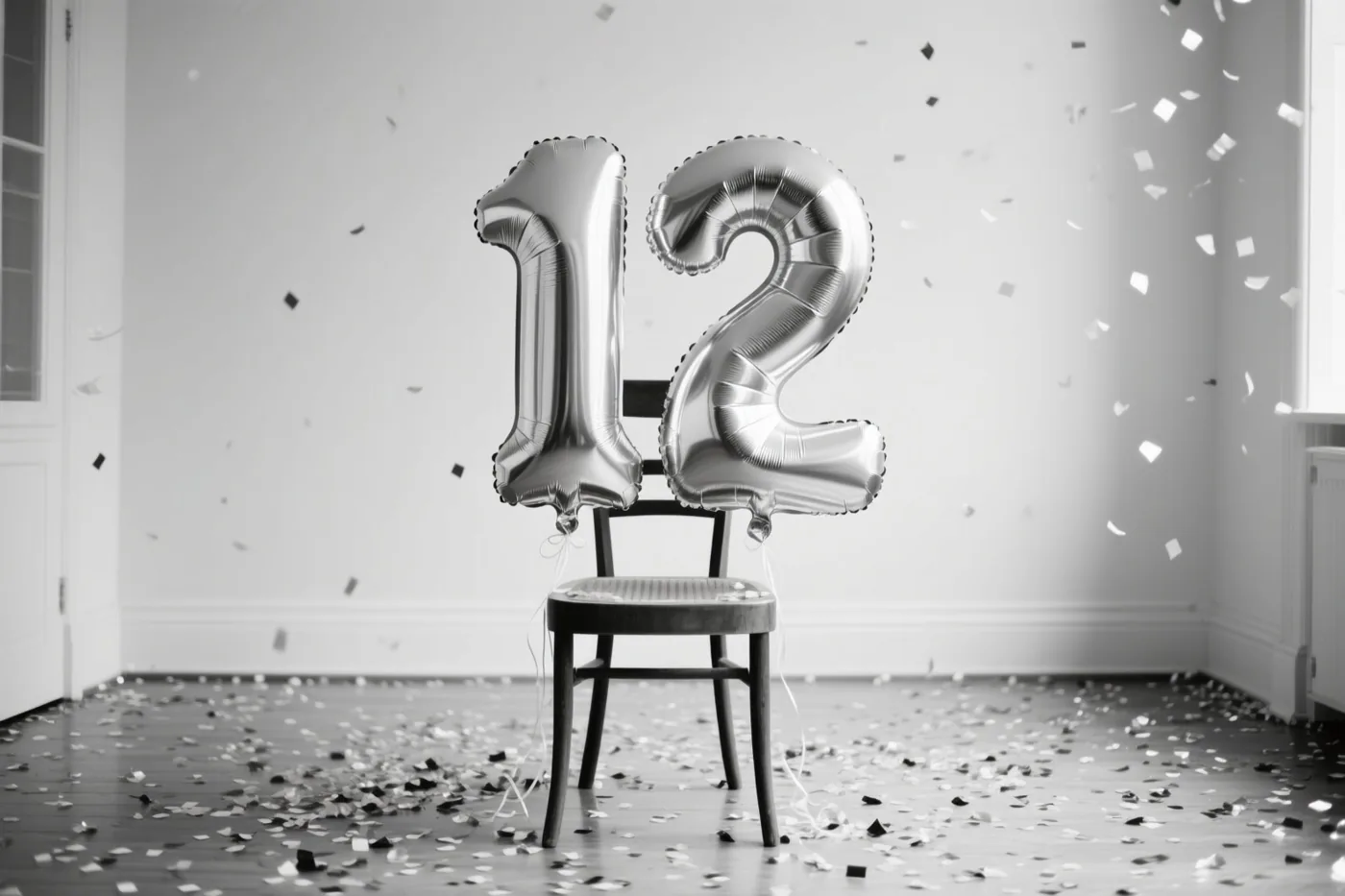 Twee grote zilverkleurige folieballonnen vormen 12 aan een houten stoel in een lichte, sober ingerichte kamer, met confetti op de vloer en in de lucht.