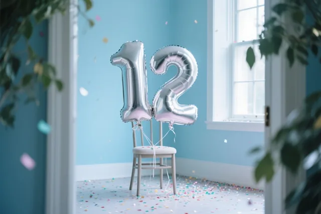 Twee zilverkleurige cijferballonnen vormen 12 aan een witte stoel in een lichte kamer met lichtblauwe muren en confetti op de vloer, met groene bladeren op de voorgrond.