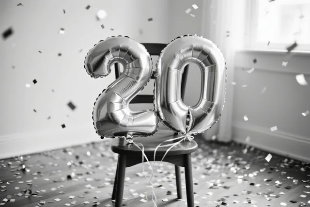 Twee grote zilveren folieballonnen vormen 20 en zijn vastgemaakt aan een houten stoel in een lichte kamer, met confetti op de vloer en in de lucht.