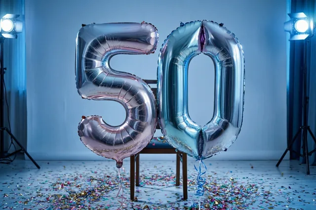 Twee grote zilveren folieballonnen in de vorm van 50 hangen aan een stoel in een studio met kleurrijke confetti op de vloer en felblauwe verlichting.