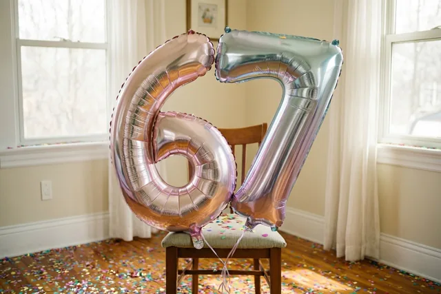 Twee grote folieballonnen in de vorm van 6 en 7, met een zilveren en pastelroze kleurverloop, hangen aan een stoel in een lichte kamer met confetti op de houten vloer.