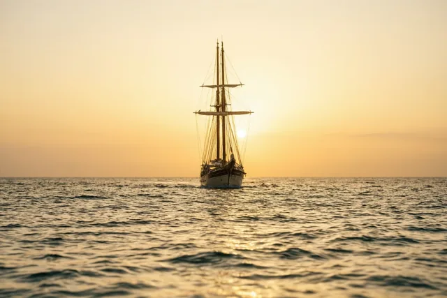 Een zeilschip vormt een silhouet op zee bij zonsondergang, met de zon achter de masten in goud en oranje tinten.