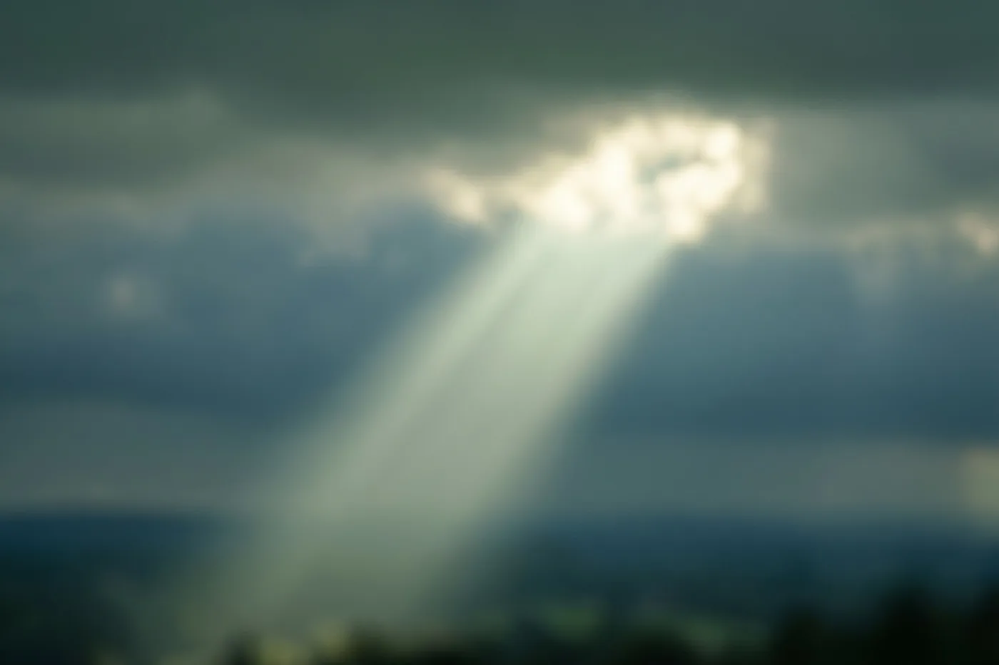 Zonnestralen vallen door donkere wolken naar beneden in een lucht met zachte blauwe, grijze en witte tinten.