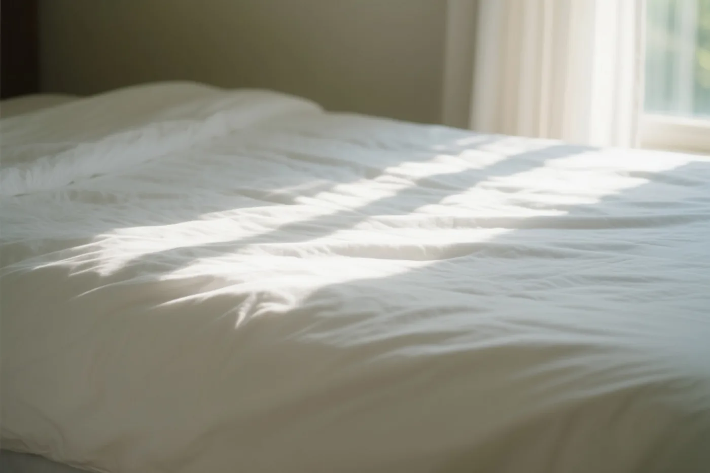 Zonlicht vormt heldere patronen op een witte sprei in een slaapkamer met een raam en gordijnen op de achtergrond.
