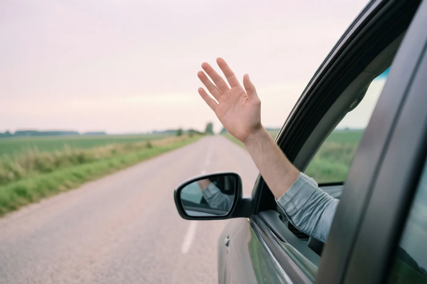 Een hand zwaait uit een autoraam op een landelijke weg met groene velden onder een bleke hemel.