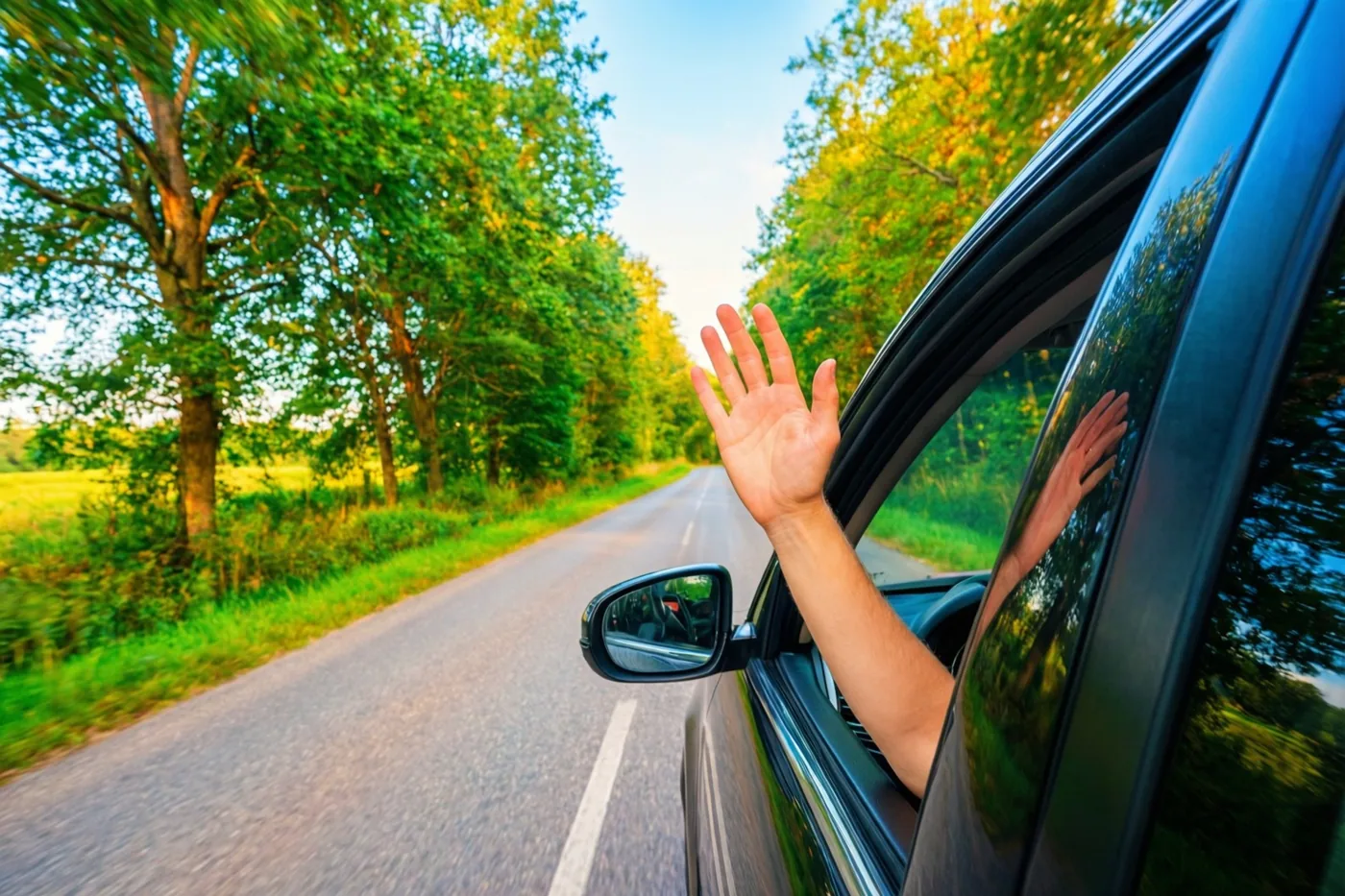 Een hand zwaait uit een autoraam op een kronkelende landweg met groene bomen in warm zonlicht.