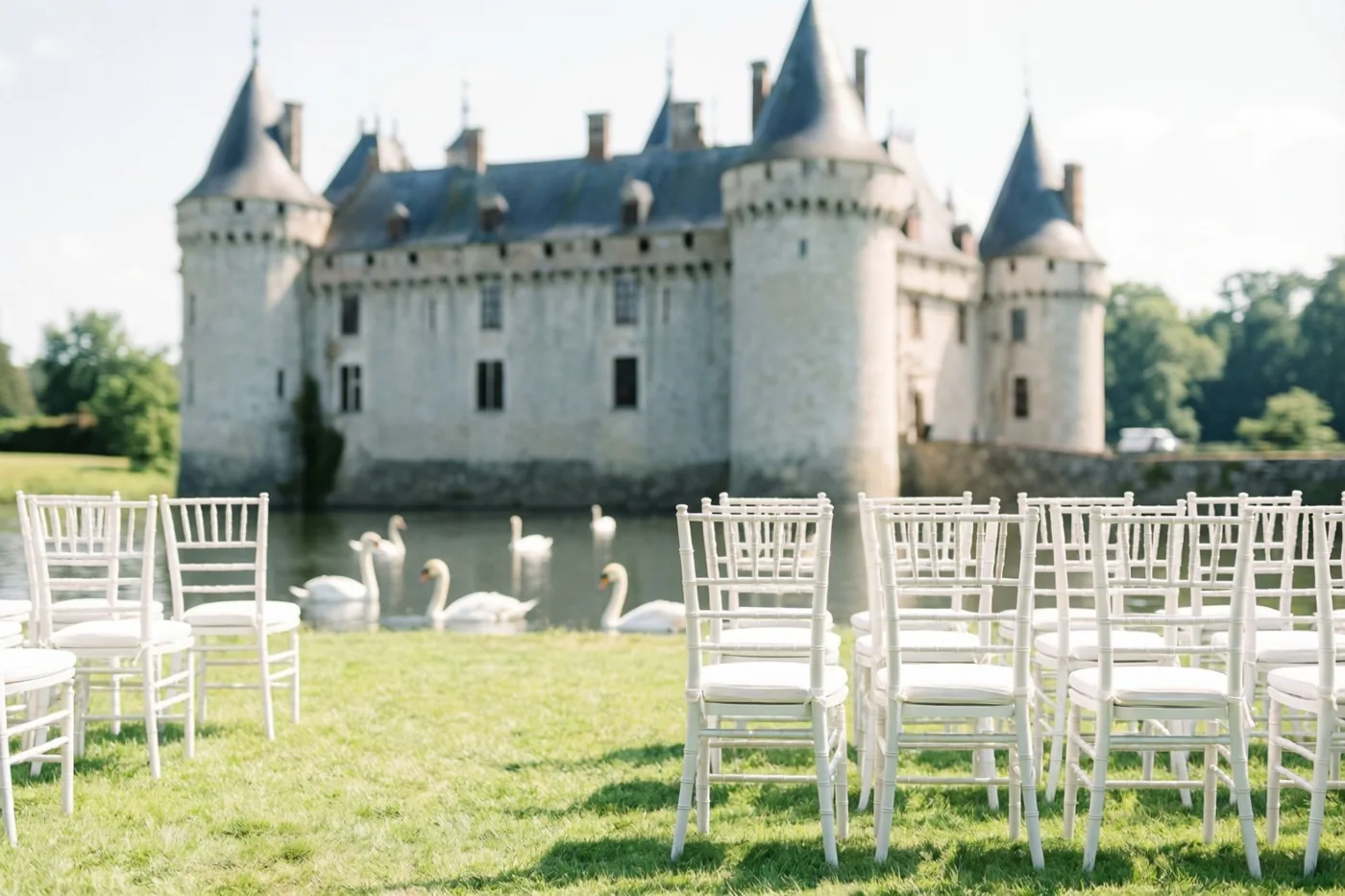 Witte stoelen staan op een groen gazon voor een kasteel met een slotgracht waarin zwanen zwemmen, in zacht daglicht.