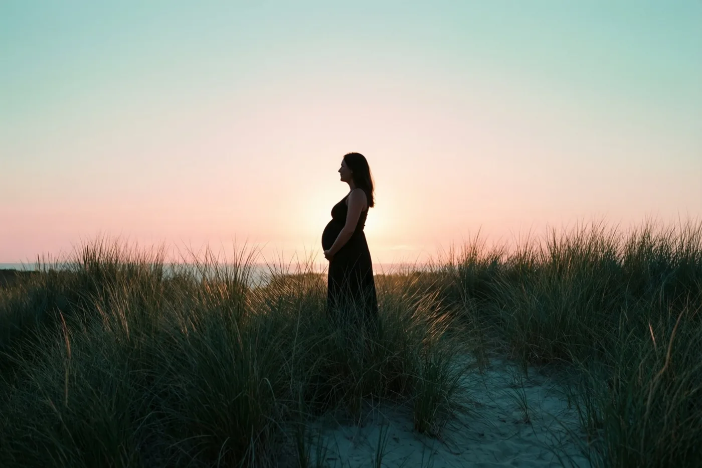 Het silhouet van een zwangere vrouw staat van opzij en op afstand in hoog duingras bij zonsopgang, in roze, perzikkleurige en blauwgroene pasteltinten.