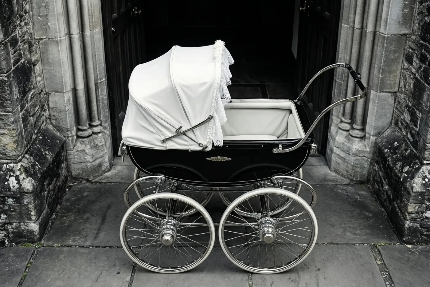 Een oude zwarte kinderwagen met chromen wielen en een wit doopmutsje staat op stenen bestrating voor een donkere kerkdeur, weergegeven in contrastrijk zwart-wit.