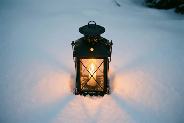 Een kleine zwarte lantaarn met een brandende kaars staat in diepe, verse sneeuw in een zwak verlicht winterlandschap met koele tinten.