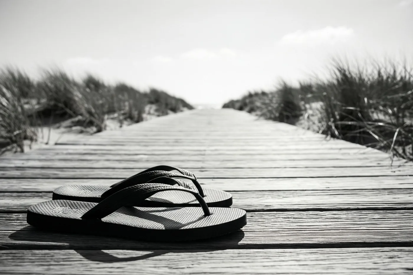 Een zwart-witfoto toont zwarte teenslippers op een houten vlonderpad dat door zandduinen loopt.