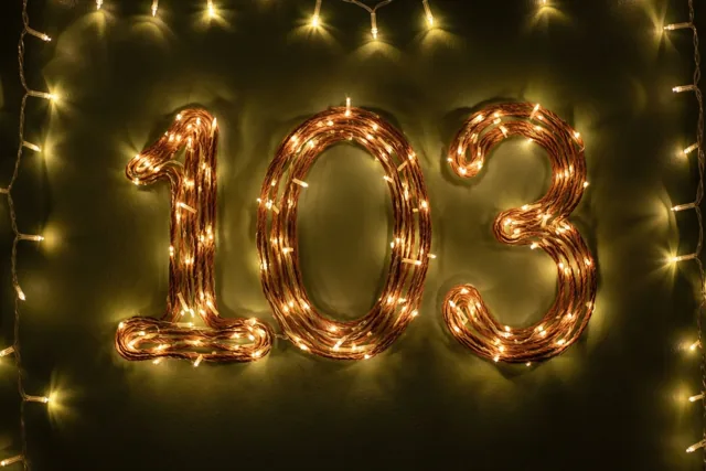 Warme lichtslingers vormen 103 op een donkere muur, met extra lichtslingers langs de randen en onscherpe lichtvlekken eromheen.