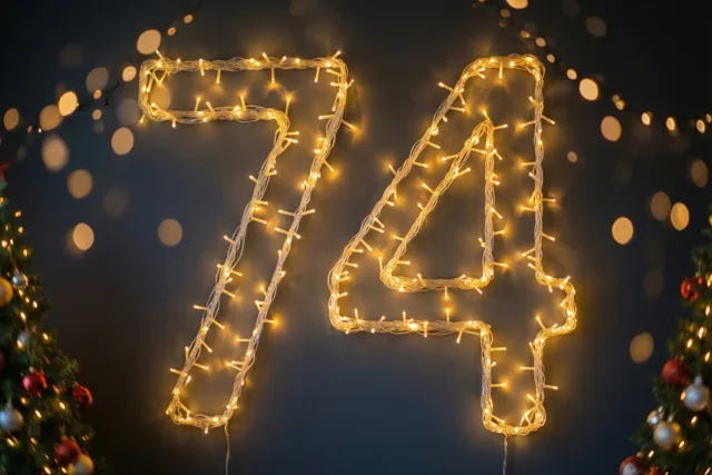 Warme lichtslingers vormen 74 op een donkere muur, met onscherpe lichtjes op de achtergrond en deels zichtbare versierde kerstbomen aan de zijkanten.