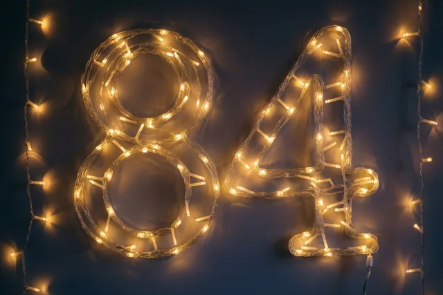 Het getal 84 bestaat uit warme lichtjes tegen een donkere muur met zachte, onscherpe lichtvlekken op de achtergrond.
