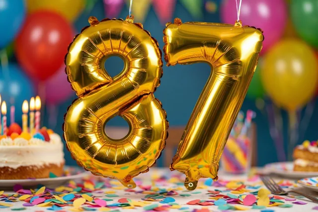 Gouden cijferballonnen vormen 87 boven een tafel met kleurrijke confetti, met daarachter een taart met brandende kaarsjes en wazige feestversiering.