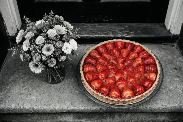 Een aardbeientaart en een klein boeket staan op een stenen drempel, waarbij de rode aardbeien de enige felle kleur vormen in een grotendeels zwart-witbeeld.