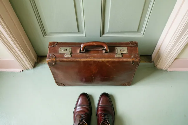 Een bruine leren retroaktetas met messing sluitingen staat op een lichtgroene vloer voor een bleekgroene paneeldeur, met gepoetste bordeauxrode schoenen ervoor.