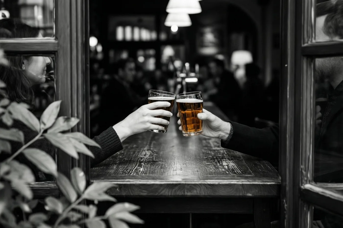 Door een raam zijn twee handen te zien die glazen amberkleurig bier heffen aan een houten cafétoog in een verder zwart-witbeeld.