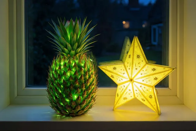 Een lichtgevende groene lantaarn in de vorm van een ananas staat 's nachts naast een gele sterlantaarn op een vensterbank.