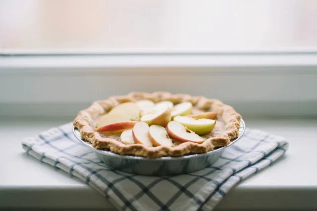 Een zelfgebakken appeltaart met appelschijfjes bovenop koelt af op een vensterbank op een blauw-wit geruite keukenhanddoek.