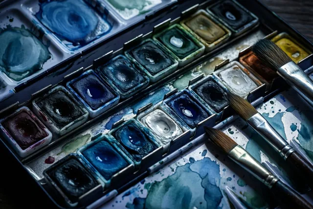 Van bovenaf is een aquarelpalet te zien met natte penselen en mengelingen van blauw, groen en aardetinten.