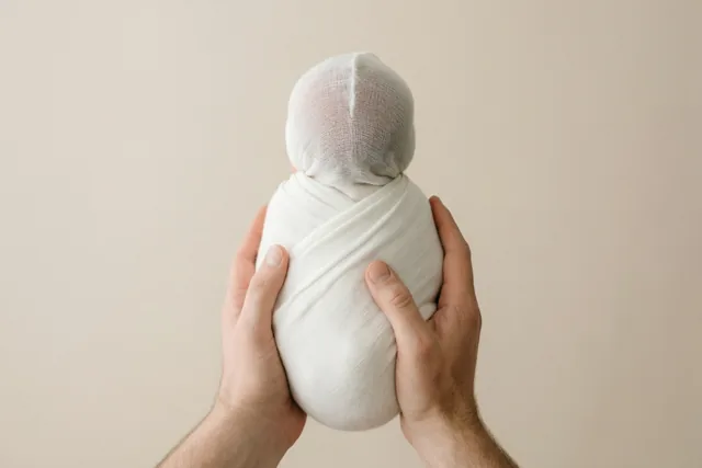 Twee handen houden voorzichtig een baby in een witte doek vast, van achteren gezien tegen een effen beige achtergrond.
