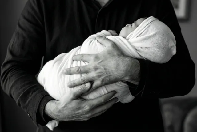Op een zwart-witfoto houden de handen van een meter of peter een baby in een witte sjaal vast, zonder zichtbare gezichten.