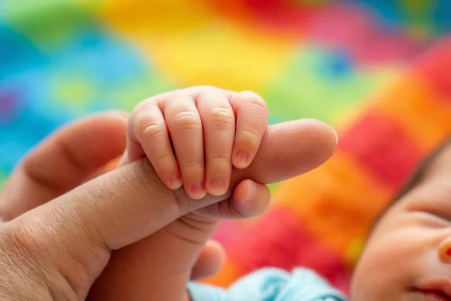 Een klein handje van een pasgeboren baby grijpt een vinger van een volwassene vast, met een onscherpe achtergrond in regenboogkleuren.