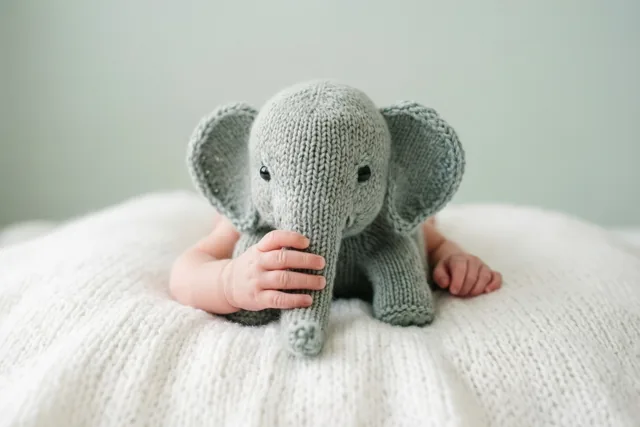 Een babyhandje houdt de slurf van een kleine grijze gebreide speelgoedolifant vast op een witte gebreide deken tegen een bleekgroene achtergrond.