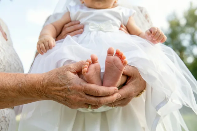 De handen van een oudere persoon houden de voetjes van een baby in een wit doopkleed vast, buiten in helder daglicht.