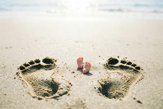 Twee kleine babyvoetjes staan in nat zand tussen twee grotere voetafdrukken van volwassenen, met de zee op de achtergrond in zacht, warm zonlicht.