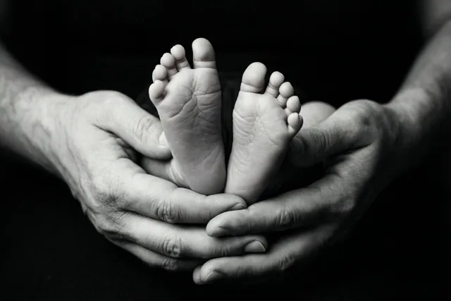 Op een zwart-witfoto ondersteunen volwassen handen de kleine voetjes van een pasgeboren baby tegen een donkere achtergrond.