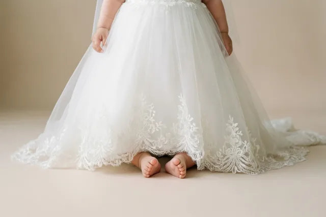 Een detailfoto toont kleine babyvoetjes onder de lange kanten zoom van een wit doopkleed, tegen een effen zachtbeige achtergrond.