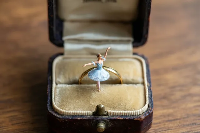 Een klein ballerinafiguurtje in een lichtblauwe tutu staat sierlijk op een gouden trouwring in een open vintage ringdoosje op een houten ondergrond in warm licht.