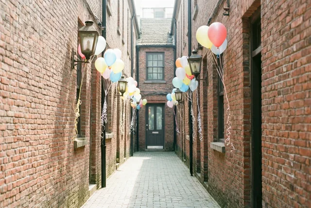 Een smal steegje met rode bakstenen muren en lichtgrijze bestrating is in zacht daglicht versierd met kleurrijke ballonnen aan oude wandlantaarns en eindigt bij een donkere voordeur.