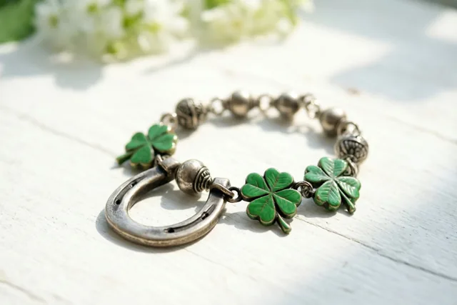 Een bedelarmband in oude stijl met een hoefijzer en groene klavertjesvier ligt op wit hout in zacht zonlicht, met onscherpe witte bloemen op de achtergrond.